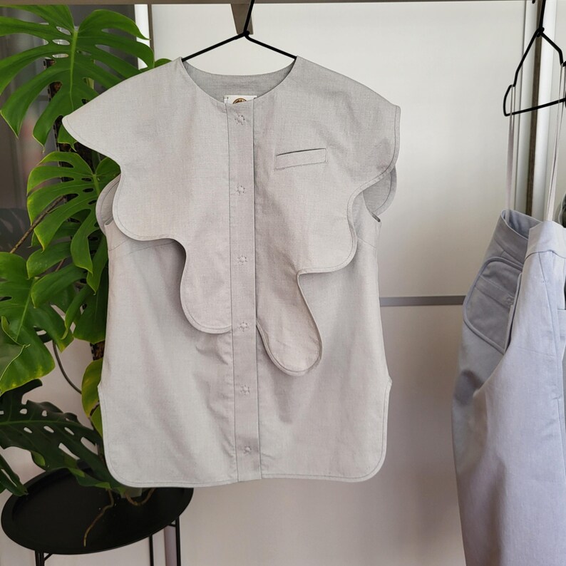 May include: A light gray sleeveless top with a unique, wavy collar design. The top has a button-up front, a small chest pocket, and is hanging on a black hanger. Another similar top is visible on the right.