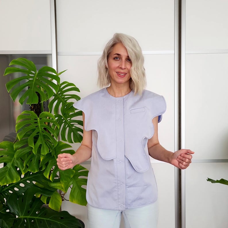 May include: A light purple button-down shirt with a unique, layered design. The shirt features a small pocket on the chest and a distinctive, asymmetrical collar. The sleeves are short, and the fabric appears to be a lightweight cotton or linen blend.