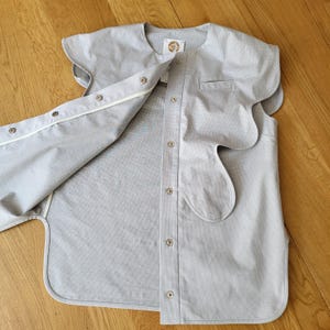 May include: A light gray button-up shirt with a unique, scalloped design on the sleeves and front. The shirt has a small pocket and is partially unbuttoned, revealing the interior. The fabric appears to be a soft, woven material.