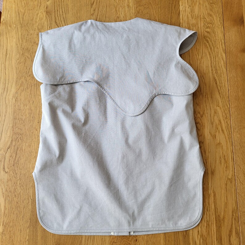 May include: A light gray bib-style garment with a unique, wavy collar design. The fabric appears to be a lightweight cotton or linen, and the garment has a simple, functional design. The bib is laid flat on a wooden surface.