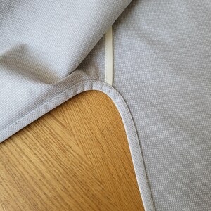 May include: A light gray textile with a subtle woven pattern. The fabric is folded over, revealing a cream-colored seam and a rounded corner. The texture appears soft and the overall aesthetic is minimalist and neutral.