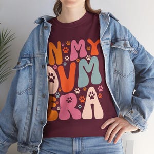 May include: A maroon t-shirt with the text "In My Own Era" in colorful, retro-style letters. The letters are surrounded by paw prints.