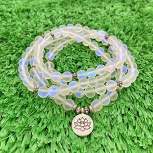 May include: A layered necklace made of iridescent, frosted glass beads. The necklace has a silver-colored lotus flower pendant. The beads are strung together with silver-colored spacers. The necklace is displayed on a green, textured surface.
