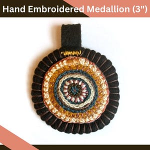 May include: Hand-embroidered medallion, approximately 3 inches in diameter, featuring concentric circles of various colors. The center has a spiral design. The outer edge is black with a looped texture, and a black strap is attached at the top.