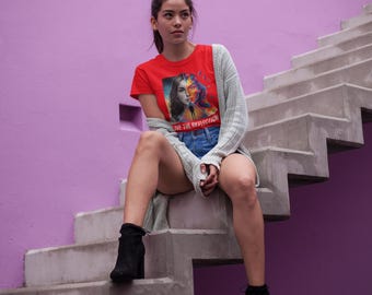 May include: A red t-shirt featuring a graphic design of a woman's face split into two halves, with the text "LIVE THE REFLECTION" in white. The person is wearing denim shorts, a light gray cardigan, and black ankle boots.