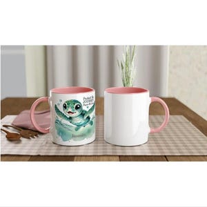 Może przedstawiać: Dwa ceramiczne kubki o pojemności 310 ml z r&oacute;żowymi brzegami. Jeden kubek przedstawia zielonego ż&oacute;łwia morskiego z tekstem "Chroń podr&oacute;ż, chroń morze" i imieniem "Jasmin". Drugi kubek jest pusty.
