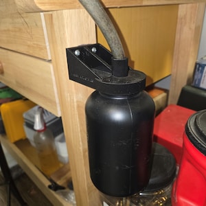 May include: A black plastic bottle with a black plastic cap and a black plastic tube attached to the cap. The bottle is mounted to a wooden surface with a black plastic bracket. The bottle is sitting on top of a clear glass jar filled with small metal objects.