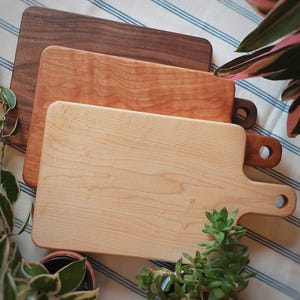 May include: Three wooden cutting boards of different sizes and wood types. The largest board is made of light wood and has a handle. The middle board is made of a darker wood and the smallest board is made of a reddish wood.