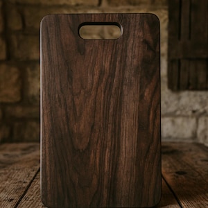 May include: A rectangular wooden cutting board with a dark brown finish. The board has a handle cut out at the top and is displayed on a wooden surface. The wood grain is visible, adding to its natural aesthetic.