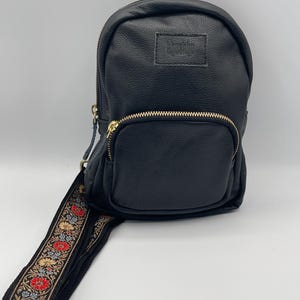 May include: A black pebbled leather sling backpack featuring a front zippered pocket with gold-tone hardware and a rectangular patch label. The adjustable strap is decorated with a woven floral pattern in red, blue, and gold thread.