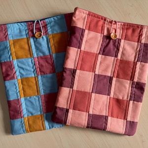 May include: Two quilted fabric cases with a patchwork design. One case features squares of blue, orange, and burgundy. The other case has a pattern of red, pink, and burgundy squares. Each case has a button closure.