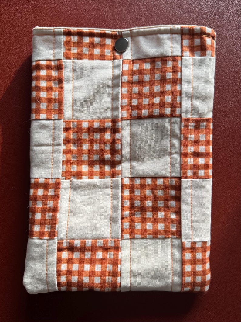 Puede incluir: Una funda de tela acolchada con un dise&ntilde;o de patchwork. La funda presenta un patr&oacute;n de cuadrados de cuadros blancos y naranjas alternados, cosidos con hilo blanco. Se ve un cierre de bot&oacute;n a presi&oacute;n plateado en la parte superior.