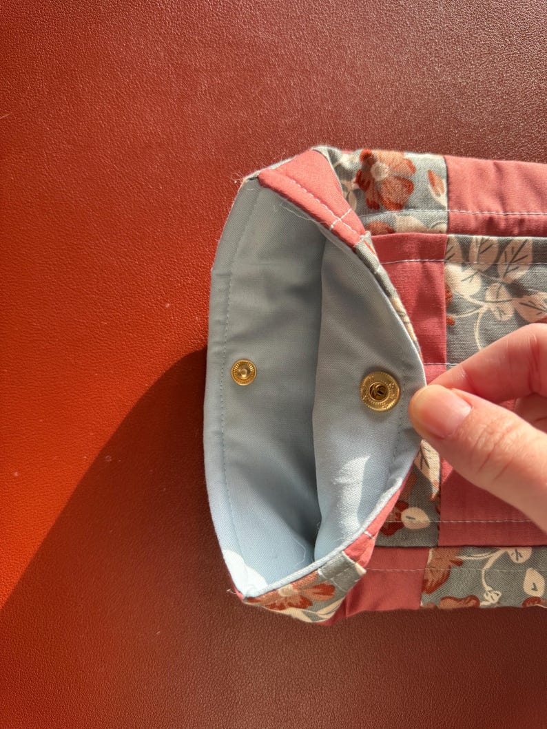 Puede incluir: Una bolsa de tela acolchada con un estampado floral en tonos rosa, azul y gris. El interior es de tela azul claro. La bolsa se asegura con dos cierres de presi&oacute;n dorados.