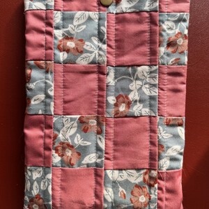 Puede incluir: Una funda de tela acolchada con un dise&ntilde;o de patchwork. La funda presenta cuadrados alternos de tela rosa polvoriento y floral con un fondo gris y flores rojas. Se ve un cierre de bot&oacute;n a presi&oacute;n de color dorado en la parte superior.