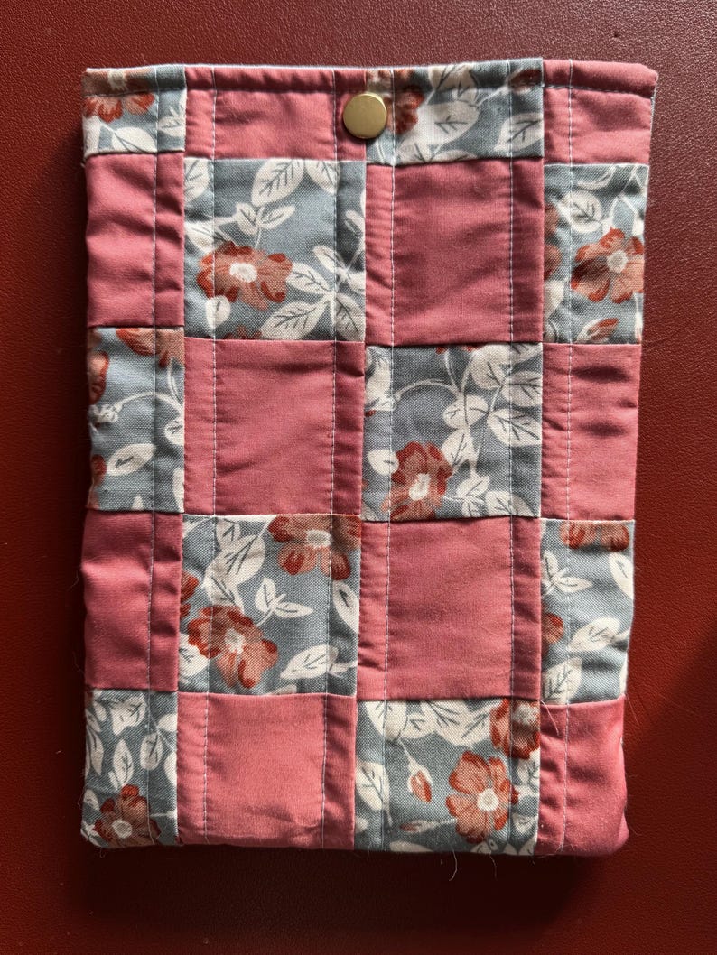 Puede incluir: Una funda de tela acolchada con un dise&ntilde;o de patchwork. La funda presenta cuadrados alternos de tela rosa polvoriento y floral con un fondo gris y flores rojas. Se ve un cierre de bot&oacute;n a presi&oacute;n de color dorado en la parte superior.