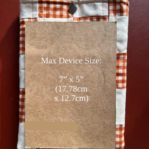 Puede incluir: Una funda de tela acolchada con un estampado de cuadros vichy naranja y blanco, asegurada con un cierre de bot&oacute;n a presi&oacute;n. Contiene un rect&aacute;ngulo marr&oacute;n con el texto "Max Device Size: 7" x 5" (17.78cm x 12.7cm)".
