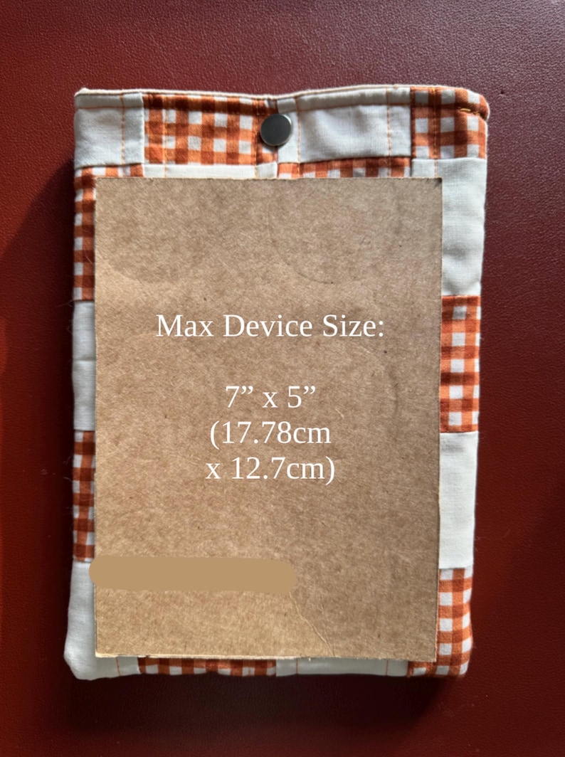 Puede incluir: Una funda de tela acolchada con un estampado de cuadros vichy naranja y blanco, asegurada con un cierre de bot&oacute;n a presi&oacute;n. Contiene un rect&aacute;ngulo marr&oacute;n con el texto "Max Device Size: 7" x 5" (17.78cm x 12.7cm)".