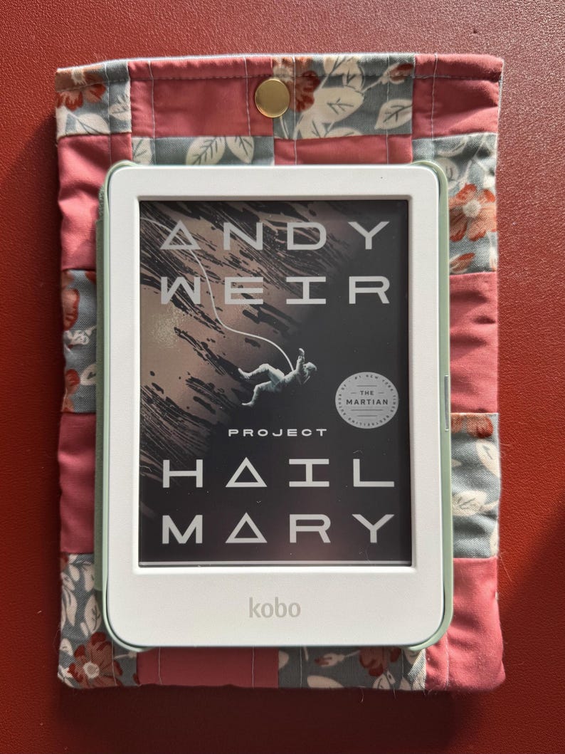 Puede incluir: Un lector electr&oacute;nico Kobo con el libro "Project Hail Mary" de Andy Weir en la pantalla, dentro de una funda acolchada. La funda es rosa y floral con un cierre de bot&oacute;n a presi&oacute;n dorado. El lector es blanco con un borde verde menta.