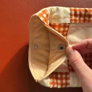 Puede incluir: Primer plano de un art&iacute;culo de tela de cuadros vichy crema y naranja con cierre de bot&oacute;n a presi&oacute;n. La tela tiene un aspecto acolchado y est&aacute; doblada, revelando un interior naranja claro. El art&iacute;culo est&aacute; sobre una superficie naranja lisa.