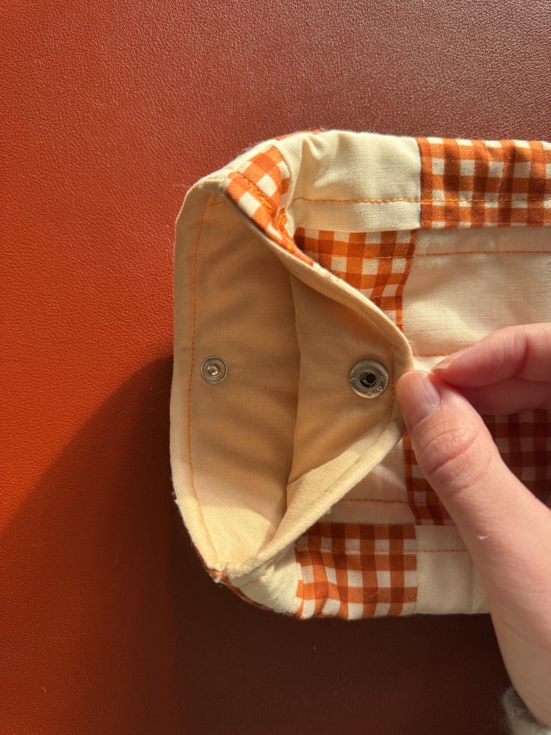 Puede incluir: Primer plano de un art&iacute;culo de tela de cuadros vichy crema y naranja con cierre de bot&oacute;n a presi&oacute;n. La tela tiene un aspecto acolchado y est&aacute; doblada, revelando un interior naranja claro. El art&iacute;culo est&aacute; sobre una superficie naranja lisa.