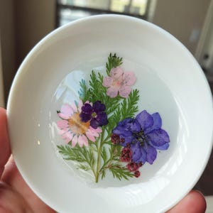 Könnte beinhalten: Eine kleine, weiße Keramikschale mit einem transparenten Harz-Innenraum. Das Harz enthält ein farbenfrohes Arrangement aus getrockneten Blumen, darunter rosa, lila und blaue Blüten, sowie grünes Laub. Die Schale ist rund und hat einen leicht erhöhten Rand.