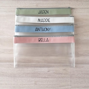 May include: Four clear zippered pouches with different colored fabric panels. The panels have the names Jaden, Maddie, Anthony, and Bella printed in black.