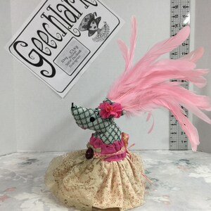 May include: A gray and white checkered fabric toy mouse wearing a pink dress with a lace overlay and a pink feather boa. The mouse has a pink flower on its head and a pink button on its dress.