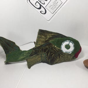 May include: A green plush fish with a white eye and a red mouth. The fish is made of different pieces of fabric sewn together.