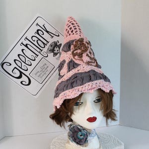 May include: A pink and gray crocheted hat with a pointed crown and a wide brim. The hat is decorated with brown crocheted embellishments. The hat is sitting on a white mannequin head with brown hair.