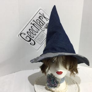 May include: A pointed witch hat made of dark blue denim fabric with a wide, grey patterned brim. The hat is displayed on a mannequin head with short brown hair, featuring a decorative fabric flower at the base and a sign reading GEECLARK.