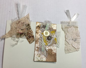 May include: Three decorative tags with a vintage aesthetic. Each tag features a different combination of lace, fabric, and buttons. The tags are attached to a white background.