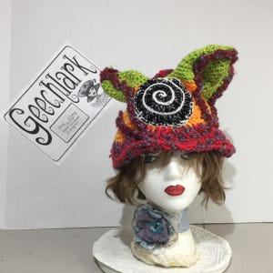 May include: A multi-colored crochet hat featuring lime green, orange, and red yarn with a prominent black and white spiral applique on the front. The hat is displayed on a mannequin head wearing a blue fabric flower accessory at the neck.