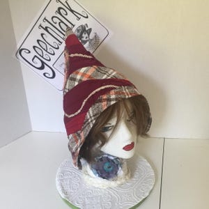 May include: A red, white, and brown plaid and quilted fabric hat with a pointed crown. The hat is on a white mannequin head with brown hair.