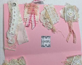 May include: A pink card with various white lace trims, pink ribbon, and a small black and white label that says "Geechlark Etsy".