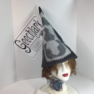 May include: A gray and white striped witch hat with a black brim and a white fabric patch with a black design. The hat is on a mannequin head with brown hair.