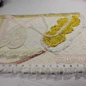 May include: A white fabric wall hanging with a variety of lace trims and a yellow crocheted piece in the upper right corner.