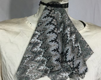 May include: A black and silver lace cravat with a floral pattern. The cravat is folded and draped over a white shirt.