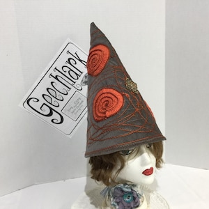 May include: A gray, cone-shaped hat with orange spiral embroidery and decorative stitching. The hat has a small, ornate gold-colored embellishment and is displayed on a mannequin head. A tag with the text "Geechlark" is visible.