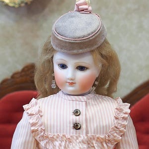 May include: A porcelain doll with blonde hair wearing a pink and white striped dress and a gray hat with a pink ribbon.