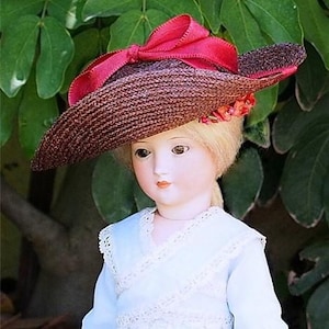 May include: A porcelain doll wearing a brown straw hat with a large red bow and floral accents. The doll is wearing a light blue dress with lace trim.