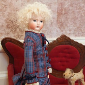 May include: A porcelain doll with blonde curly hair wearing a blue and red checked dress with a red velvet bow. The doll is standing next to a small red velvet chair.