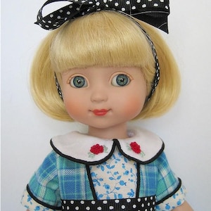Peut inclure: Une poupée aux cheveux blonds et aux yeux bleus, portant un serre-tête à pois noirs et blancs avec un nœud. Elle est vêtue d'une robe à carreaux bleus et blancs, dotée d'un col blanc brodé de fleurs rouges et d'une ceinture à pois.