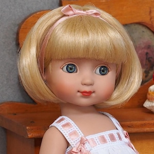Peut inclure: Une poupée aux cheveux blonds coupés au carré, portant un bandeau en ruban rose avec un petit nœud. La poupée a des yeux bleus, des joues roses et des lèvres rouges. Elle porte un vêtement blanc sans manches avec des rubans roses sur les bretelles.
