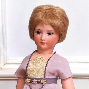 May include: A porcelain doll with a light pink dress, a gold and purple beaded necklace, and a short blonde wig.