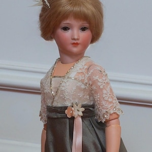May include: A porcelain doll with blonde hair, wearing a white lace top with a gray skirt and a pink sash. The doll has a delicate facial features and is wearing a small feather ornament in her hair.