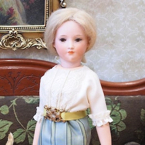 May include: A porcelain doll with blonde hair, wearing a white blouse with lace trim, a yellow belt with a decorative buckle, and a blue and white striped skirt.