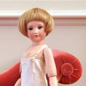 Könnte beinhalten: Eine Porzellanpuppe mit einer blonden Perücke, die ein weißes Unterkleid trägt. Die Puppe hat bemalte blaue Augen und einen rosigen Teint.