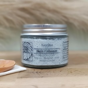 Può includere: Un piccolo barattolo di sali da bagno etichettato "Nautylus Bain Calmant" con un'etichetta grigia e bianca che presenta un disegno di conchiglia di nautilus. L'etichetta include anche il testo "Melange pour le bain, Sel de mer, Sel d'Epsom, Avoine colloidale et lavande".