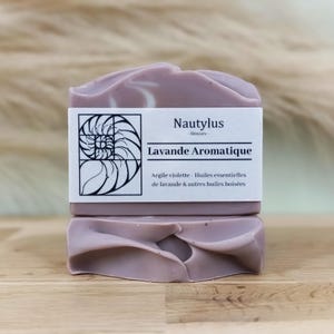 Puede incluir: Una barra de jabón con aroma a lavanda con un diseño de remolino morado y blanco. El jabón está etiquetado como "Nautylus - Skincare - Lavande Aromatique - Argile violette - Huiles essentielles de lavande & autres huiles boisées."