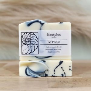 Puede incluir: Dos barras de jabón blanco con vetas negras. El jabón está etiquetado como "Nautilus Skincare Le Tonic" y enumera los ingredientes como "Hules essentielles de bergamote & lavande - charbon actif bio".